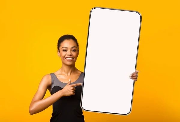 Lächelnde junge schlanke schwarze Frau zeigt mit dem Finger auf ein riesiges Smartphone mit leerem Bildschirm, isoliert auf orangefarbenem Hintergrund — Stockfoto