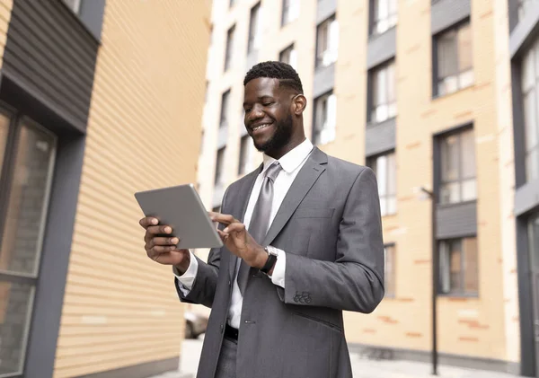 Heureux employé de l'entreprise noire qui travaille avec une tablette numérique pendant la pause déjeuner, marchant près du centre de bureau à l'extérieur — Photo