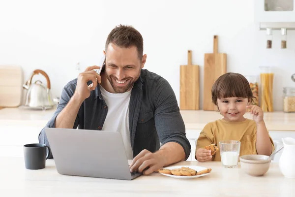 Cute little boy with glass of milk smiling to camera, sitting at kitchen with his father freelancer working from home — Stock Photo, Image