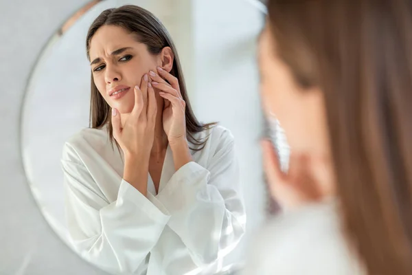 Frustrado joven mujer con problema de piel Popping grano en la mejilla cerca del espejo — Foto de Stock