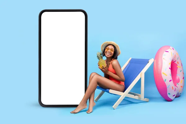 Joyful young woman in bikini sitting in lounge chair near giant smartphone with blank screen, mockup for mobile app — Stock Photo, Image