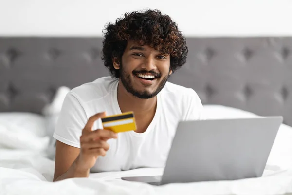 Indian guy laying in bed with credit card and laptop — Stock Photo, Image
