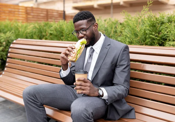 Ung afrikansk amerikansk kontorsarbetare som äter lunch utomhus, dricker kaffe och äter smörgås, sitter på bänken — Stockfoto