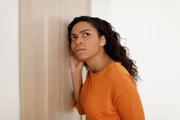 Mulher afro-americana preocupada ouvindo através de porta fechada em pé interior — Fotografia de Stock