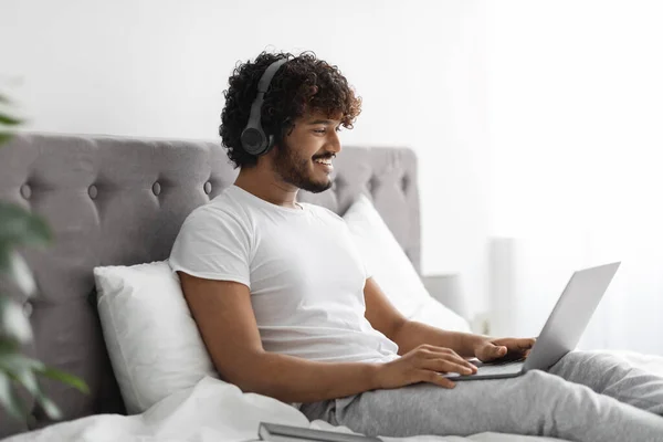 Relajado indio chico disfrutando de fin de semana en la cama, utilizando el ordenador portátil — Foto de Stock