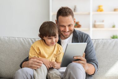 Çocuk gelişimi içeriği. Genç baba, küçük sevimli oğlana dijital tablet gösteriyor. Çocuk internette eğitim programından hoşlanıyor.