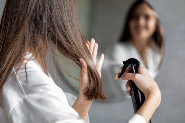 Cuidado com os cabelos. Jovem mulher aplicando spray de cabelo enquanto estava perto do espelho no banheiro — Fotografia de Stock