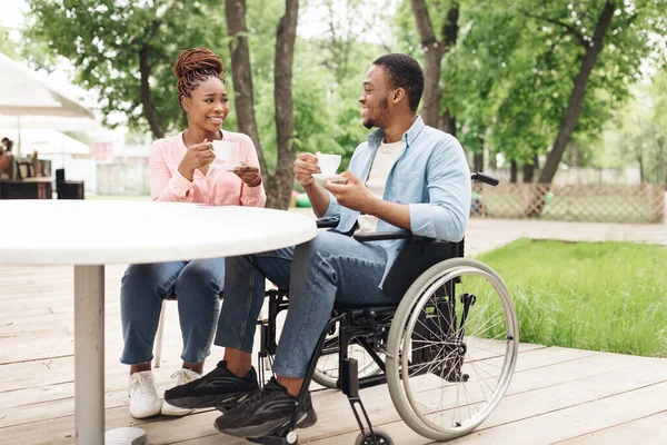 Millennial czarny facet na wózku inwalidzkim mając randkę z piękną kobietą w kawiarni na świeżym powietrzu, korzystających z kawy i rozmowy — Zdjęcie stockowe
