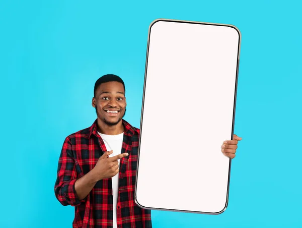 Sorprendido chico afroamericano joven feliz en puntos casuales dedo en el teléfono inteligente enorme con pantalla en blanco —  Fotos de Stock