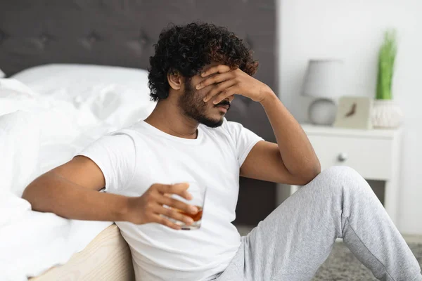 Depressed indian man drinking alcohol, sitting on floor by bed — Stock Photo, Image