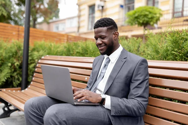 Απομακρυσμένη δουλειά έξω. Αυτοπεποίθηση Αφροαμερικανός επιχειρηματίας ή διευθυντής με κοστούμι χρησιμοποιώντας φορητό υπολογιστή, κάθεται στον πάγκο έξω — Φωτογραφία Αρχείου