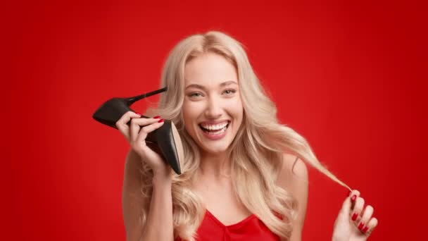 Feminino falando segurando sapatos de salto alto como telefone, fundo vermelho — Vídeo de Stock