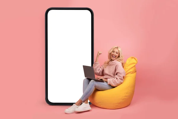 Spännande ung blond dam med bärbar dator gör JA gest, sitter i bönan väska stol nära stora smartphone med mockup — Stockfoto