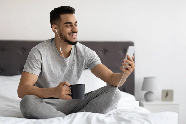 Διαδικτυακή κλήση. Θετικός νεαρός Άραβας χρησιμοποιώντας Smartphone στο κρεβάτι — Φωτογραφία Αρχείου