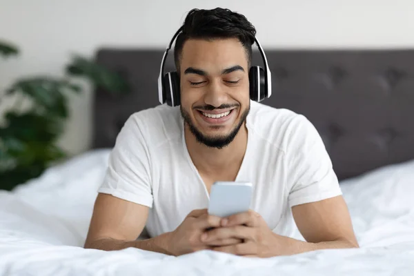 Moderne Technologien. Schöner junger Mann aus dem Nahen Osten entspannt sich im Bett mit Smartphone — Stockfoto