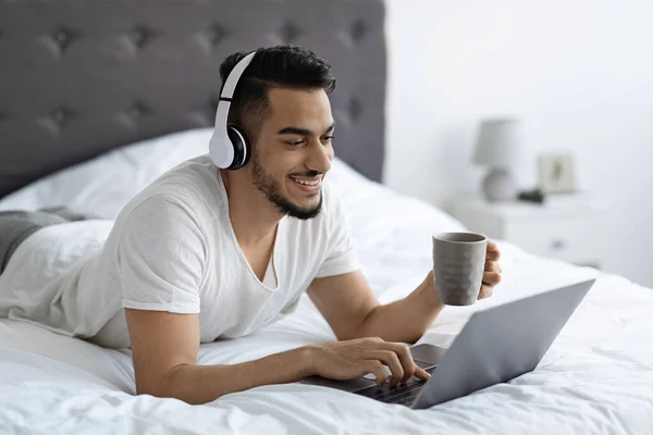 Medio oriente chico en auriculares relajante en la cama con el ordenador portátil y el café — Foto de Stock