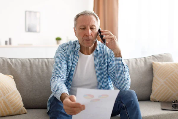 Сосредоточенный взрослый мужчина держит отчет о чтении бумаги разговаривая по телефону — стоковое фото