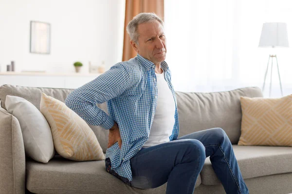 Homem maduro com dor nas costas sentado no sofá em casa — Fotografia de Stock