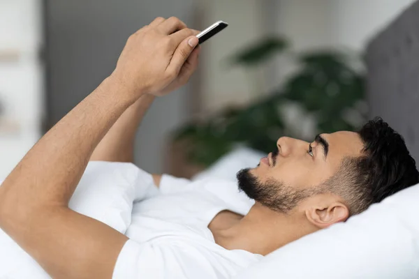 Πλάγια άποψη του νεαρού Άραβα χρησιμοποιώντας Smartphone, ενώ βρίσκεται στο κρεβάτι — Φωτογραφία Αρχείου
