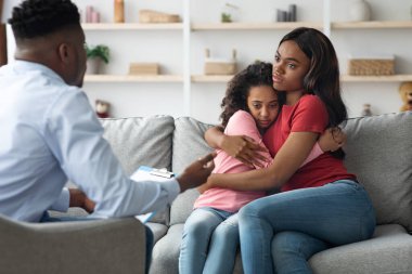 Çocuk psikoloğuna giderken korkmuş kızını kucaklayan siyahi bir kadın.