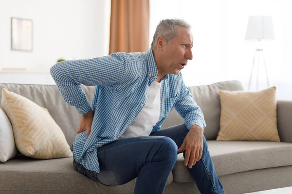 Mature man with side back pain sitting on couch at home — Stock Photo, Image