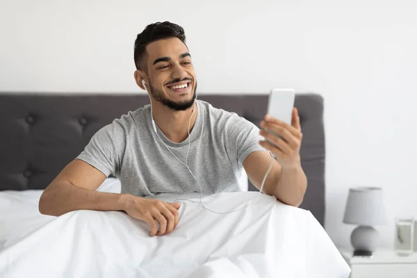 Διαδικτυακή επικοινωνία. Happy Arab Man Χρήση Smartphone Στο Κρεβάτι Για Βίντεο Κλήση — Φωτογραφία Αρχείου