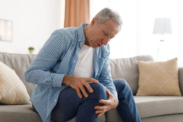 Reifer Mann mit Knieschmerzen sitzt auf Couch zu Hause — Stockfoto