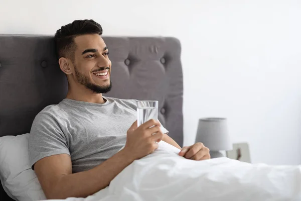 Hombre árabe guapo feliz sentado en la cama y sosteniendo el vaso con agua — Foto de Stock