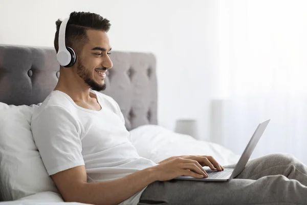 Młody Arab Man w słuchawkach bezprzewodowych Korzystanie z laptopa podczas relaksu w łóżku — Zdjęcie stockowe