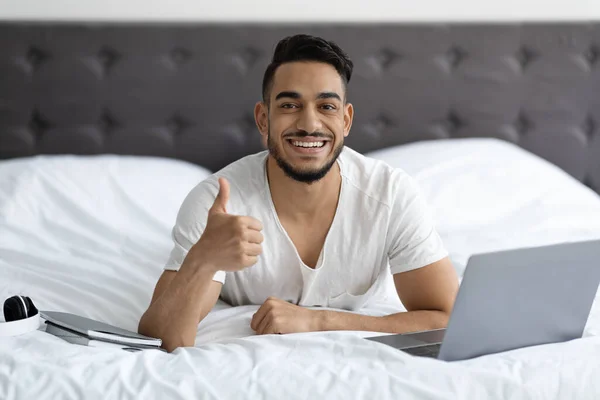 Koncepcja wolnego strzelca. Szczęśliwy Arab człowiek z laptopem w łóżku Pokazuje kciuk w górę — Zdjęcie stockowe