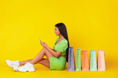 Akıllı telefon kullanan mutlu bir kadın, alışveriş torbalarıyla yerde oturuyor.