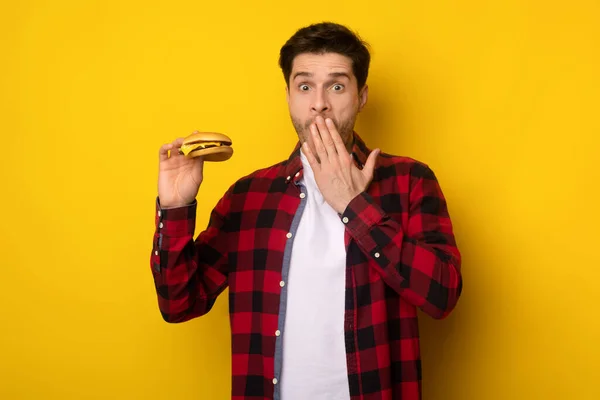 Engraçado jovem cara segurando hambúrguer e cobrir a boca — Fotografia de Stock