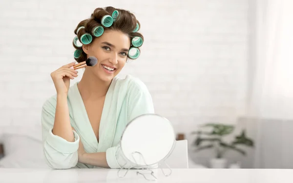 Atractiva dama haciendo maquillaje usando cepillo cosmético sentado en el dormitorio —  Fotos de Stock