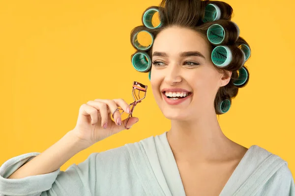 Alegre dona de casa curling cílios e cabelo posando sobre fundo amarelo — Fotografia de Stock