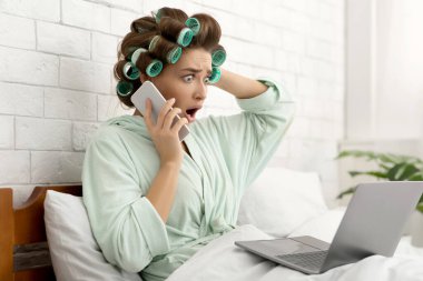 Şok olmuş ev hanımı cep telefonuyla konuşuyor. Laptop kullanıyor. Yatak odasında oturuyor.