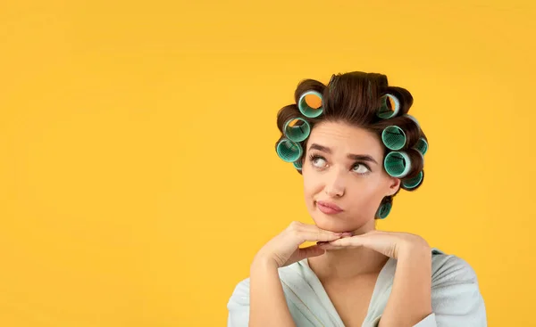 Mulher cuidadosa com encrespadores de cabelo pensando sobre fundo amarelo — Fotografia de Stock