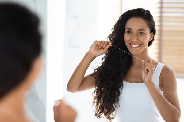 Mujer árabe sonriente usando hilo dental mientras está de pie cerca del espejo en el baño —  Fotos de Stock