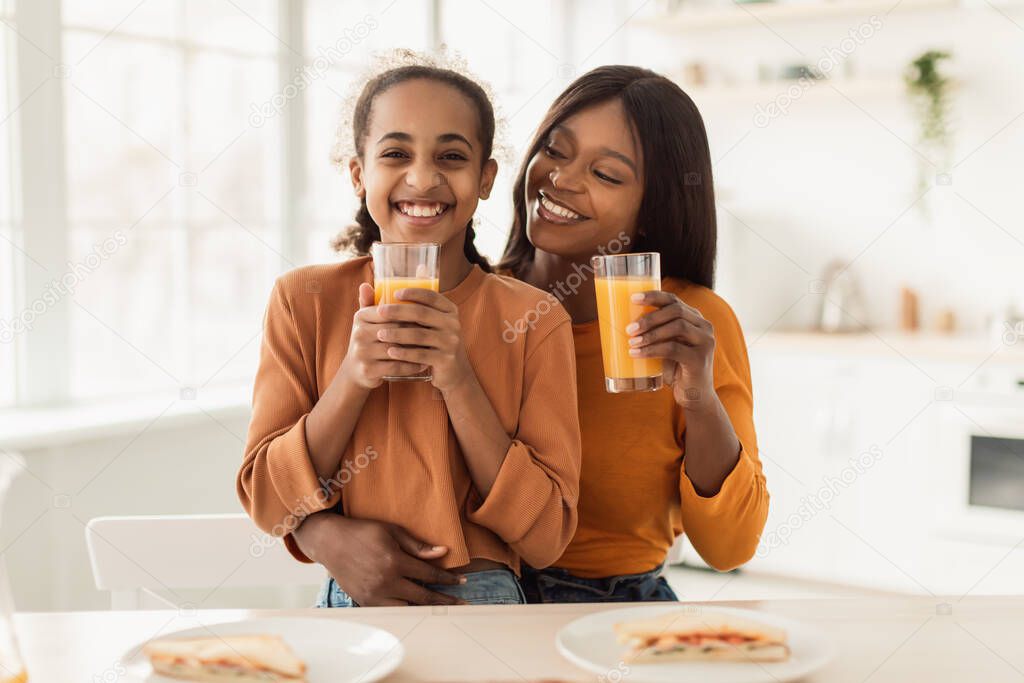 African Mom And Daughter Drinking Juice Holding Glasses In Kitchen