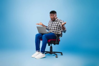 Bin yıllık siyahi adam dizüstü bilgisayarla koltukta oturuyor, mavi stüdyo arka planında online galibiyete şaşırıyor.