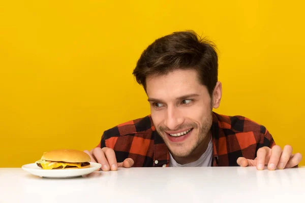 Fome jovem homem olhando para hambúrguer espreitando para fora tabela — Fotografia de Stock