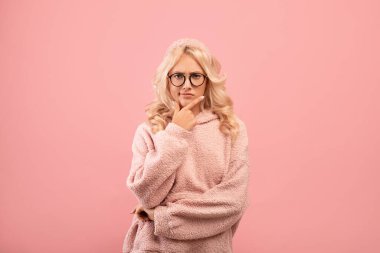 Ciddi bir kadın soru sormayı düşünüyor, çenesine dokunuyor, kameraya bakıyor, pembe arka planda duruyor.