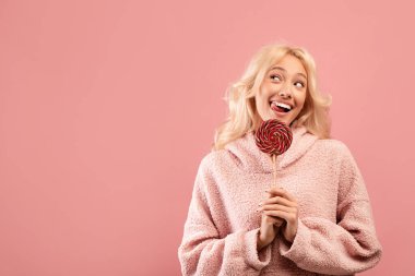 Tatlıya düşkünlük. Çubukta şeker tutan, boş alana bakan, gülümseyen, pembe arka planlı, oyuncu bir kadın.