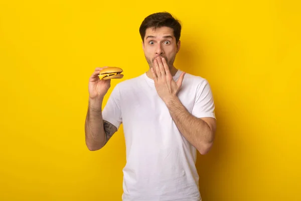 Chico divertido sosteniendo hamburguesa y cubriendo la boca — Foto de Stock