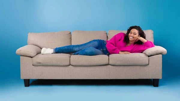 Hermosa mujer negra feliz joven en ropa casual tumbado en el sofá y sonriendo a la cámara en el fondo del estudio azul —  Fotos de Stock
