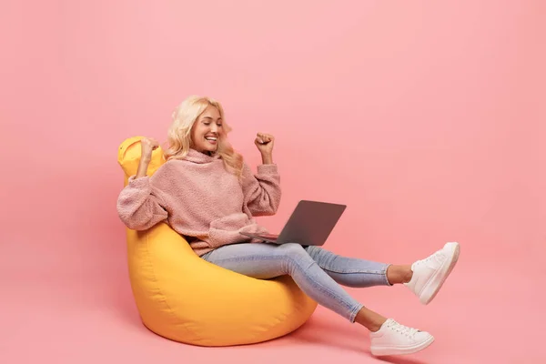 Με χαρά γυναίκα που χρησιμοποιεί το laptop και σηκώνοντας τα χέρια, διαβάζοντας σπουδαία νέα και γιορτάζοντας την επιτυχία, κάθεται στην καρέκλα beanbag — Φωτογραφία Αρχείου