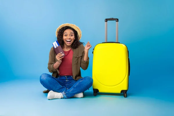 Aufgeregte junge schwarze Frau mit hellem Koffer, Pass und Flugticket sitzt im Schneidersitz und macht JA-Geste — Stockfoto