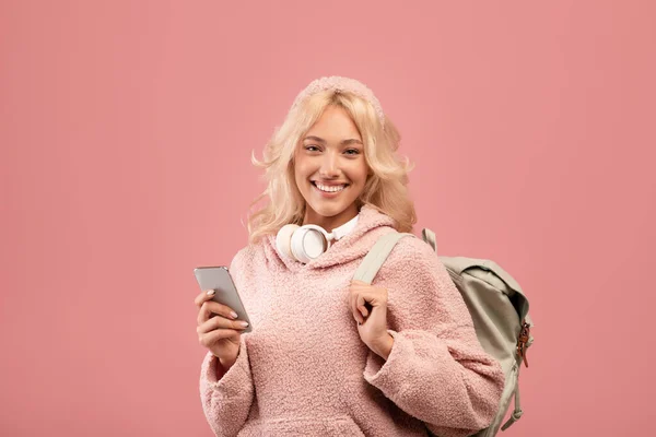 Nadšený dáma student pomocí smartphone a usmívat se na kameru, zatímco stojí s batohem přes růžové pozadí — Stock fotografie
