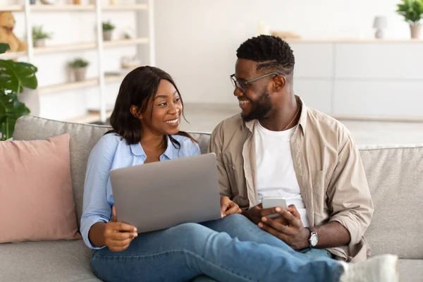 Happy black couple sitting on couch, using cellphone and pc
