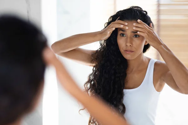 Aynaya bakan ve yüzündeki kırışıklıklara dokunan endişeli genç bir kadın. — Stok fotoğraf