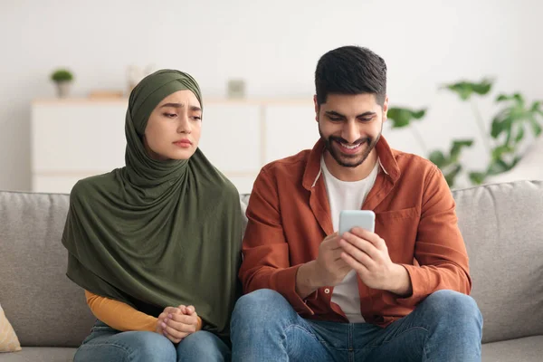 Mulher muçulmana infeliz olhando para o marido SMS usando Smartphone Interior — Fotografia de Stock